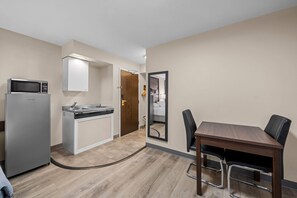 Cocina compacta con microondas, lavabo y armario, mesa de comedor con sillas y un espejo que refleja un dormitorio.