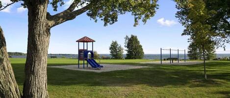Un parc avec une aire de jeux, un grand arbre et une vue sur l’eau.