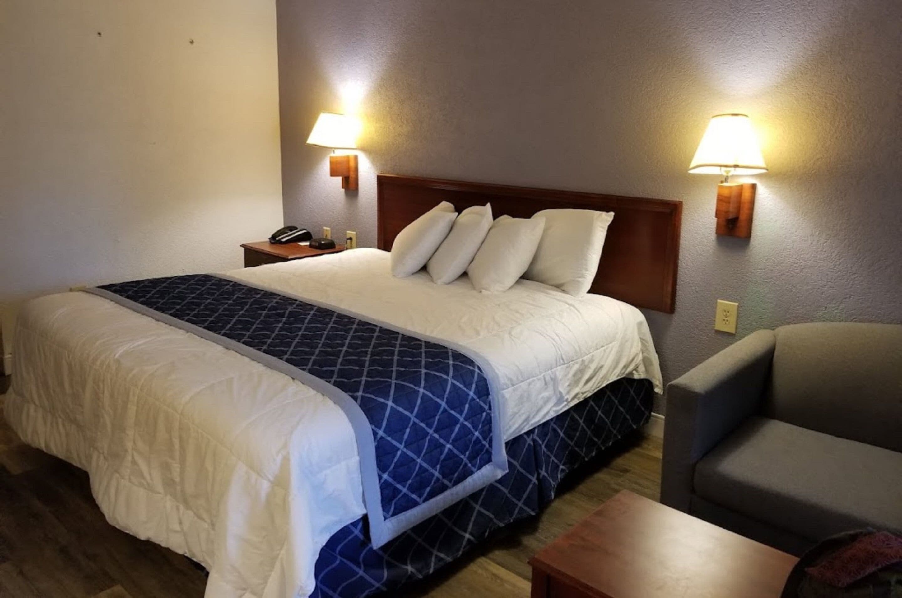 Una habitación de hotel con una cama grande, dos lámparas de noche, un cabecero de madera, un sillón gris y una pequeña mesa de madera.