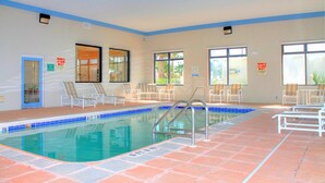 An indoor swimming pool with tiled flooring, lounge chairs, and windows with views.