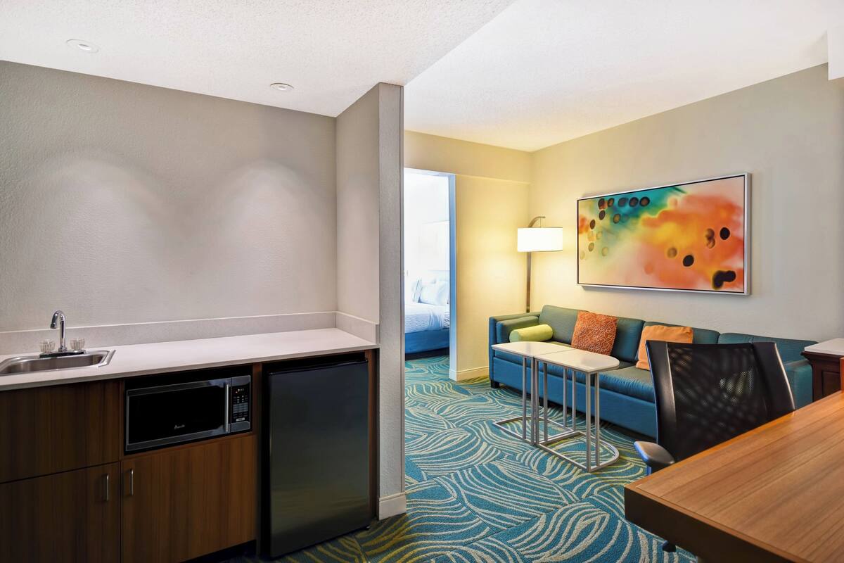 A compact hotel room with a kitchenette, a sofa, a small table, and a colorful abstract painting on the wall.