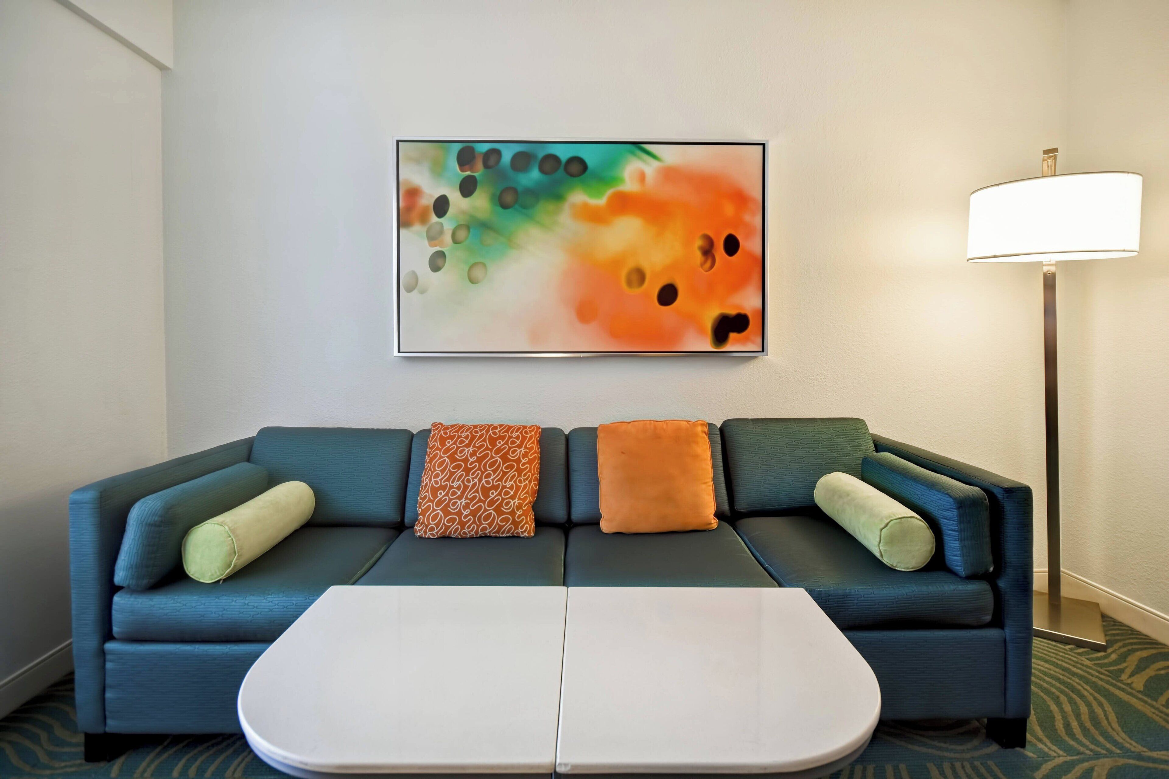 A modern hotel room with a blue sofa, two white side tables, and a colorful abstract painting on the wall.