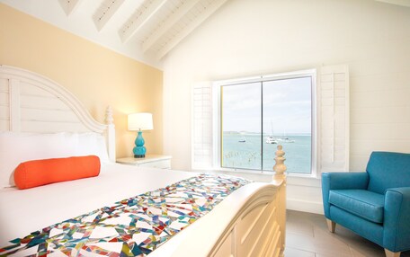 A bedroom with a bed, a window with a view of the sea, a blue armchair, and a lamp on a nightstand.
