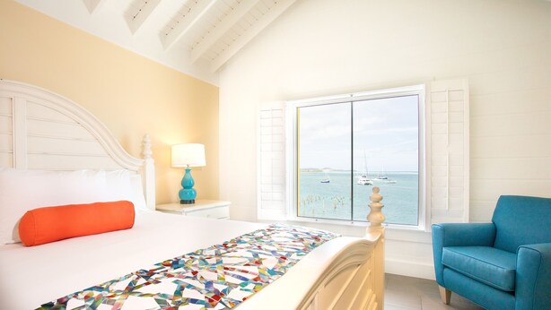Un dormitorio con cama, ventana con vista al mar, sillón azul y lámpara en una mesita de noche.