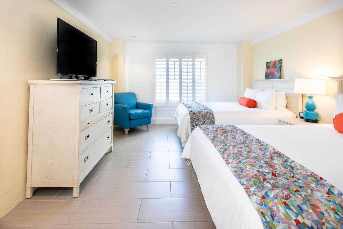 Habitación de hotel con televisión, una silla azul, dos camas con ropa de cama colorida, un aparador y una ventana con persianas.