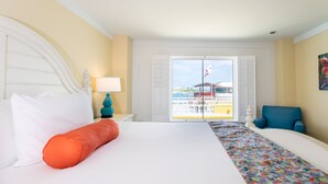 Habitación de hotel con cama, una colcha colorida, una silla azul y vista a un edificio a través de una puerta de vidrio.