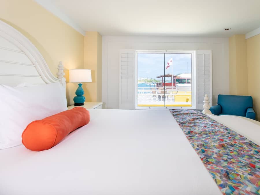 Une chambre d’hôtel avec un lit, une couvre-lit aux couleurs vives, une chaise bleue et une vue sur un immeuble par une porte vitrée.