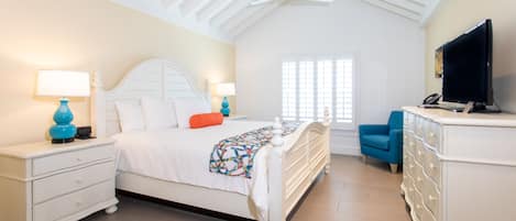 Un dormitorio con una cama blanca, dos mesitas de noche con lámparas, un aparador, un televisor y una ventana con persianas.