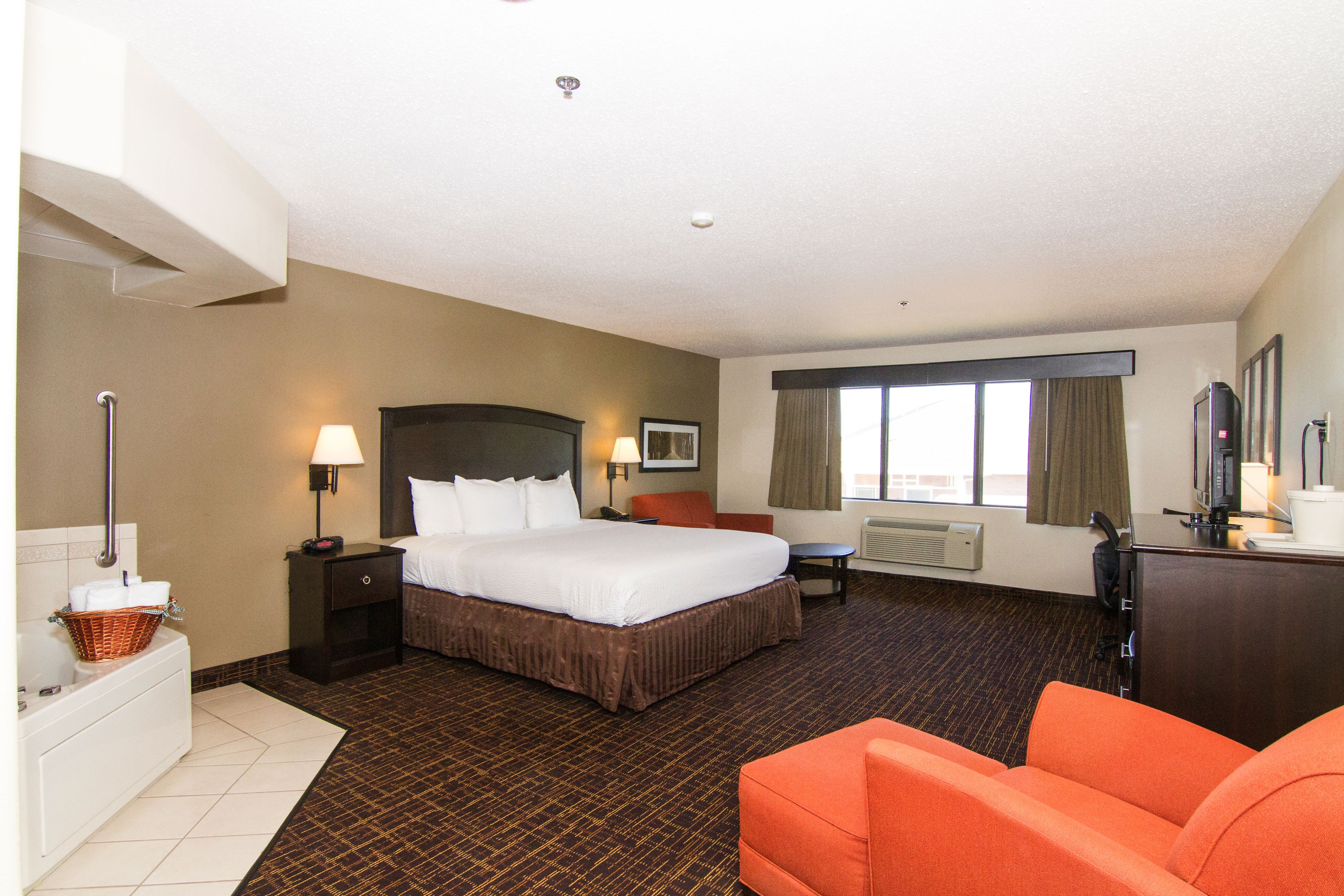 A hotel room with a large bed, a desk, an orange chair, a desk lamp, a TV, and a window with curtains.