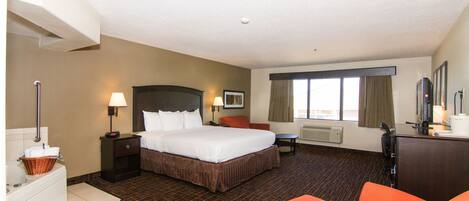 A hotel room with a large bed, a desk, an orange chair, a desk lamp, a TV, and a window with curtains.