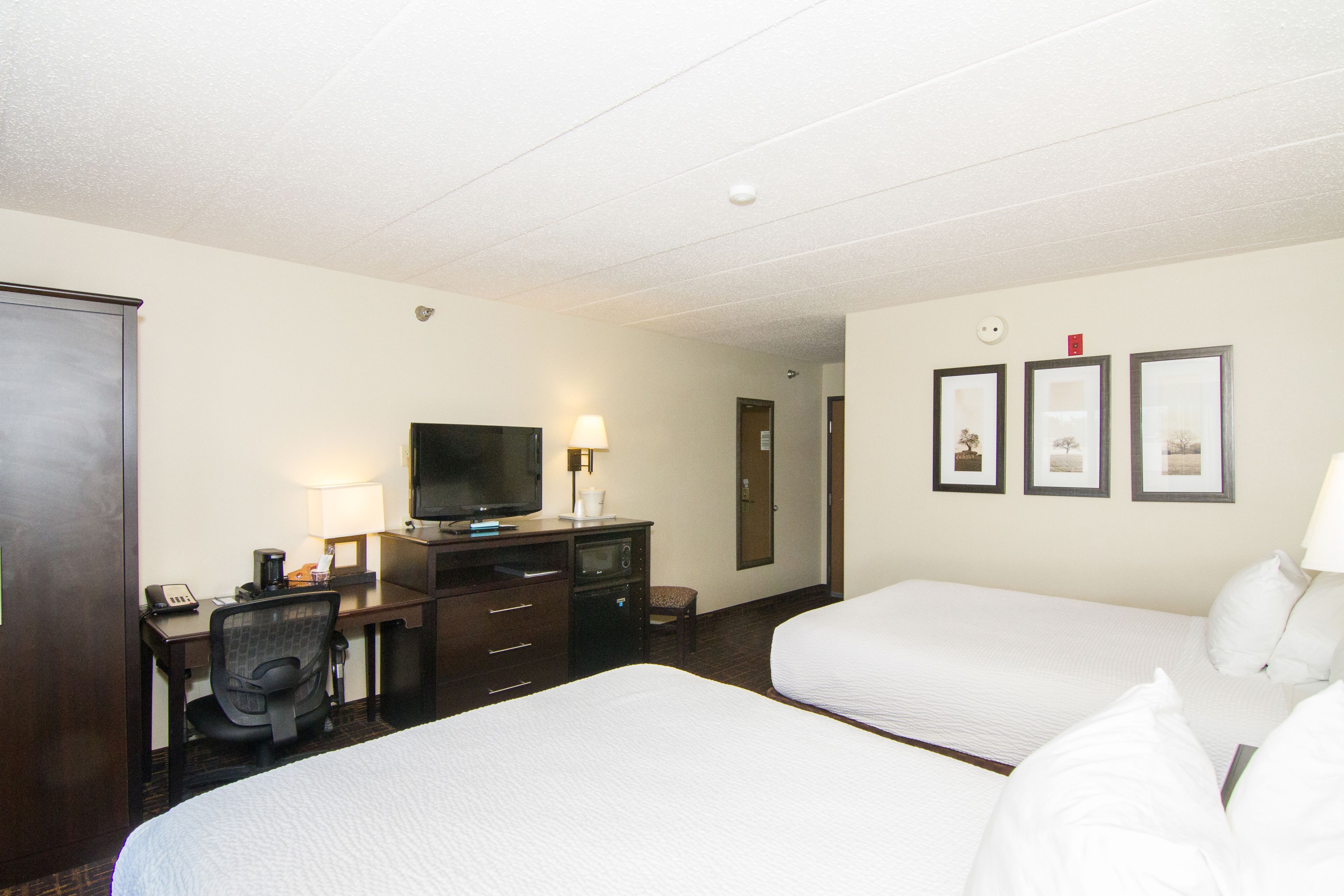 A hotel room with two beds, a desk with a television, a chair, and a small table with a lamp.