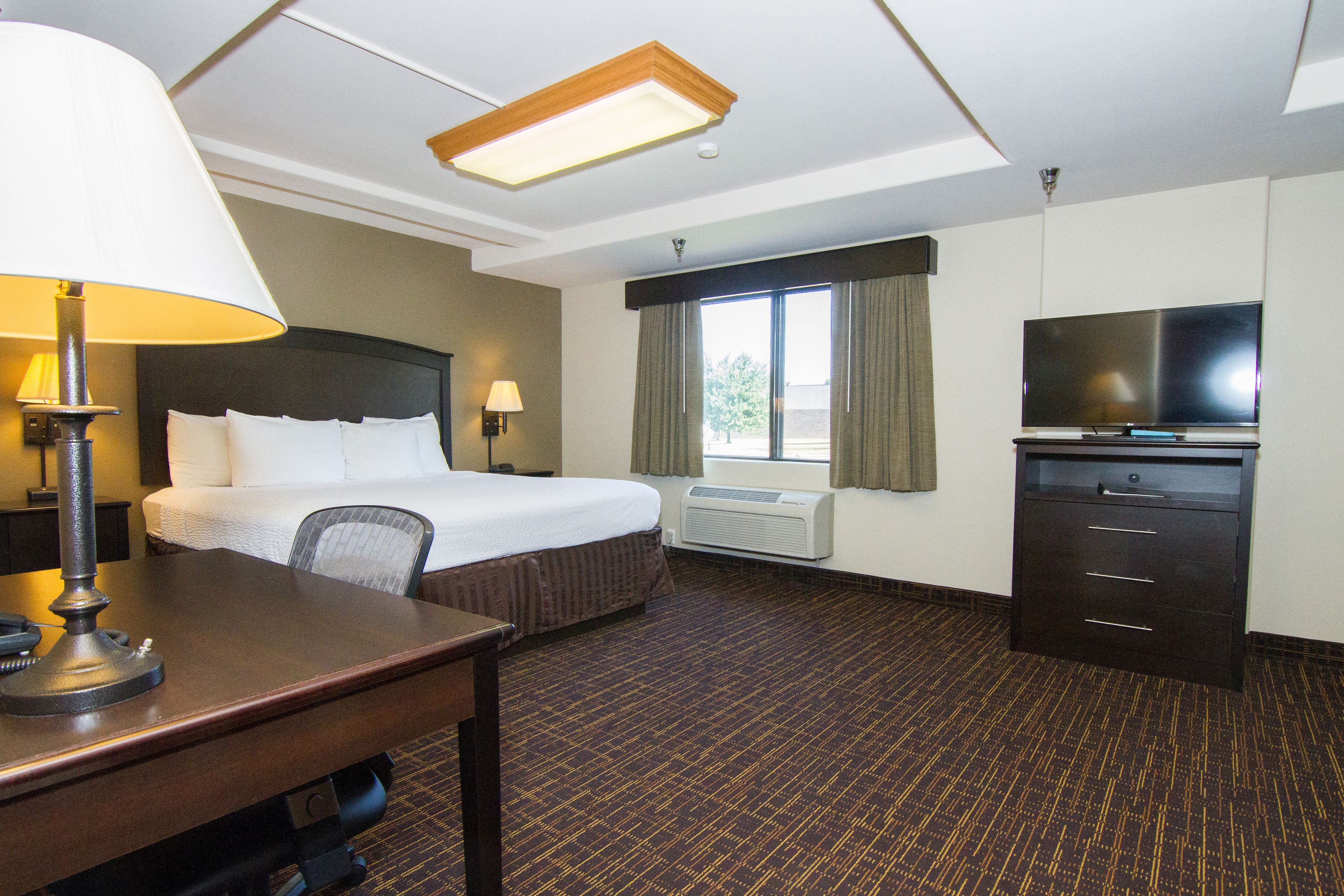 A hotel room with a bed, desk, lamp, TV, and air conditioning unit.