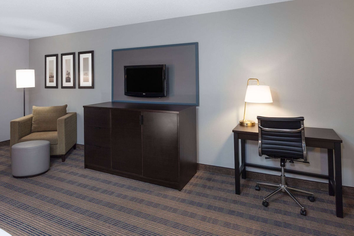 A hotel room with a flat-screen TV mounted on the wall, a desk with a lamp, a cushioned chair, a wardrobe, and a round ottoman.