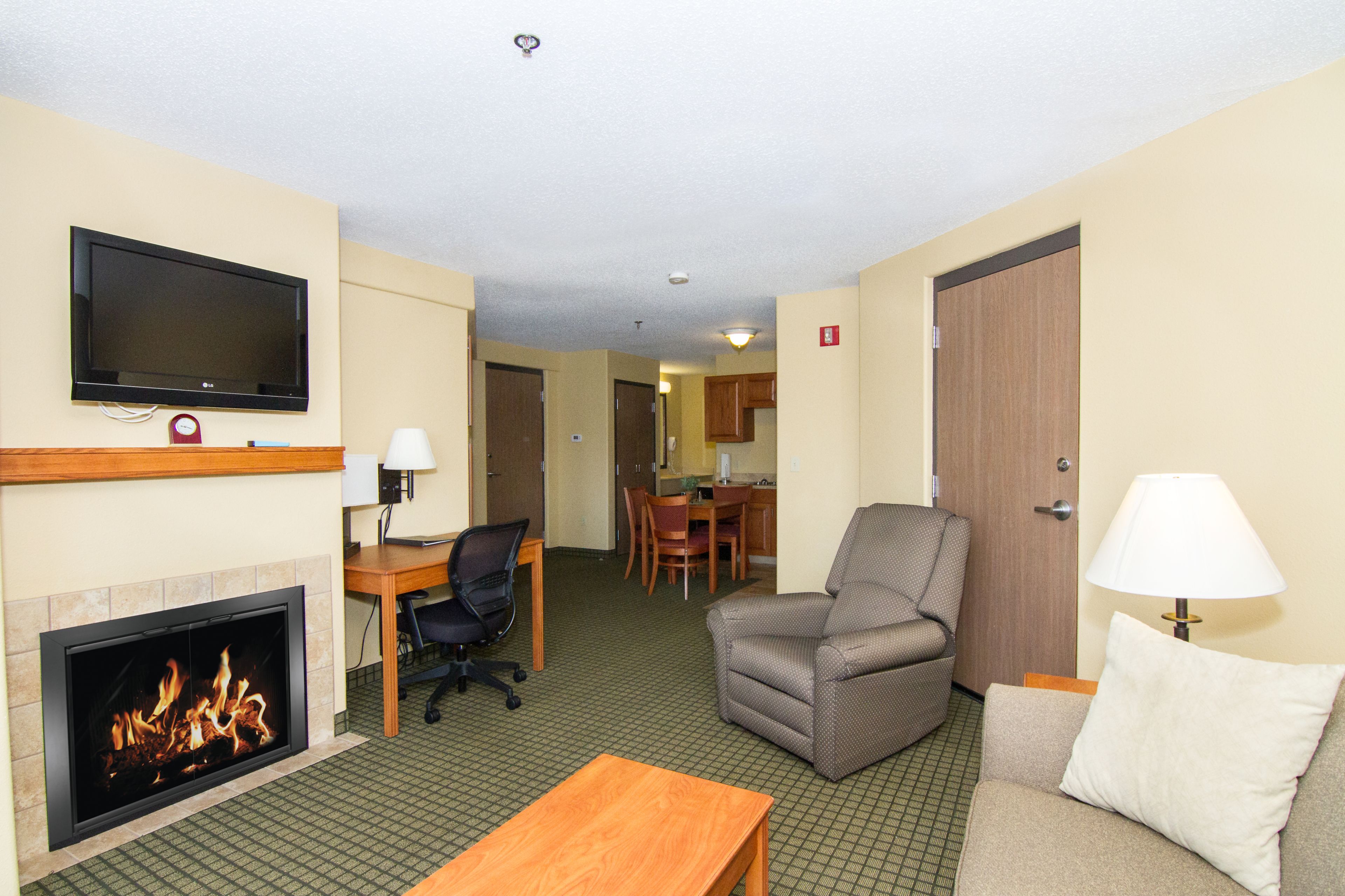 A hotel room with a flat-screen TV mounted above a fireplace, a sofa, a small table, and a dining area in the background.