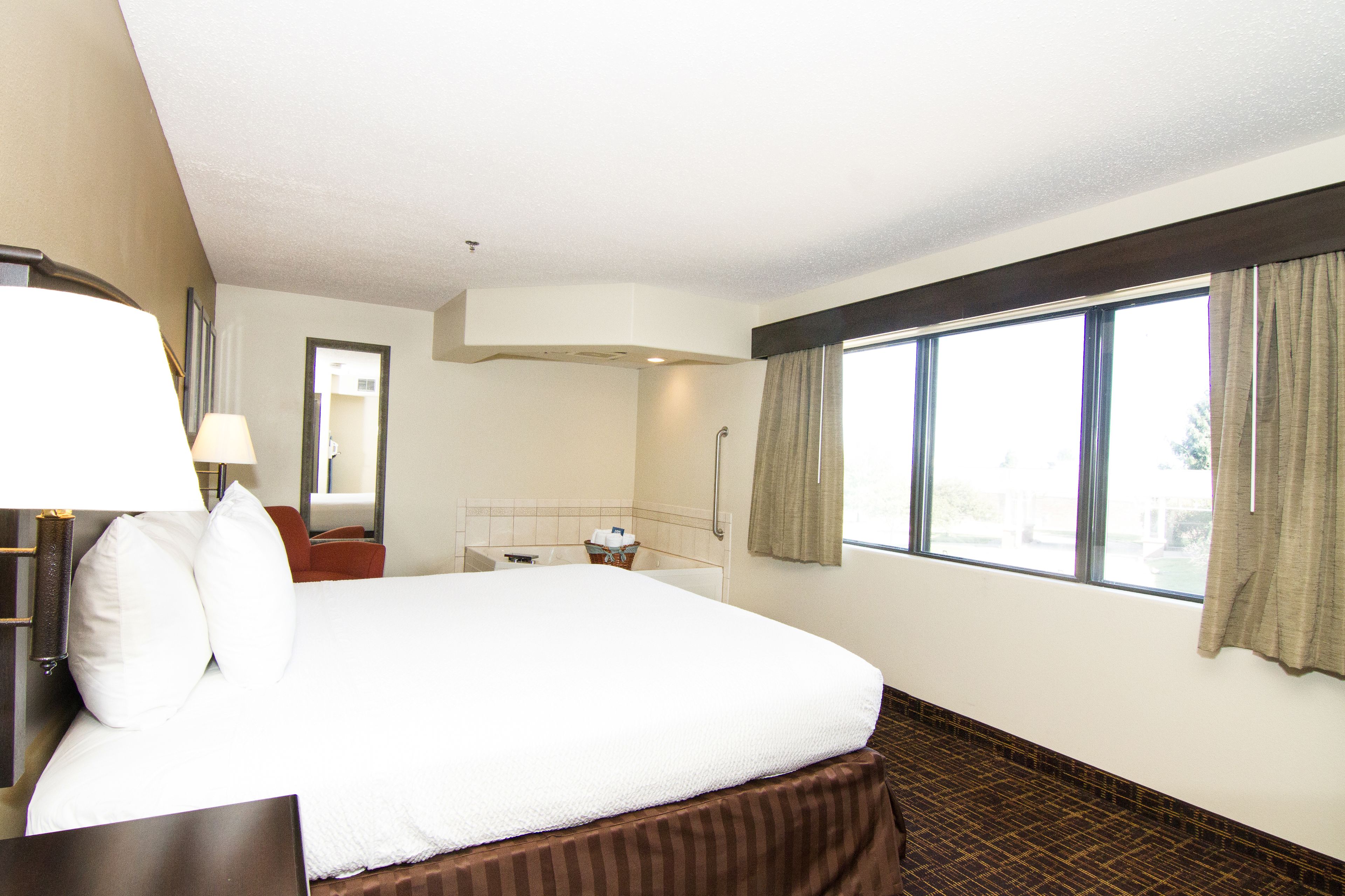 A hotel room with a large bed, a bedside table with a lamp, a bathroom with a shower, and a window with curtains.