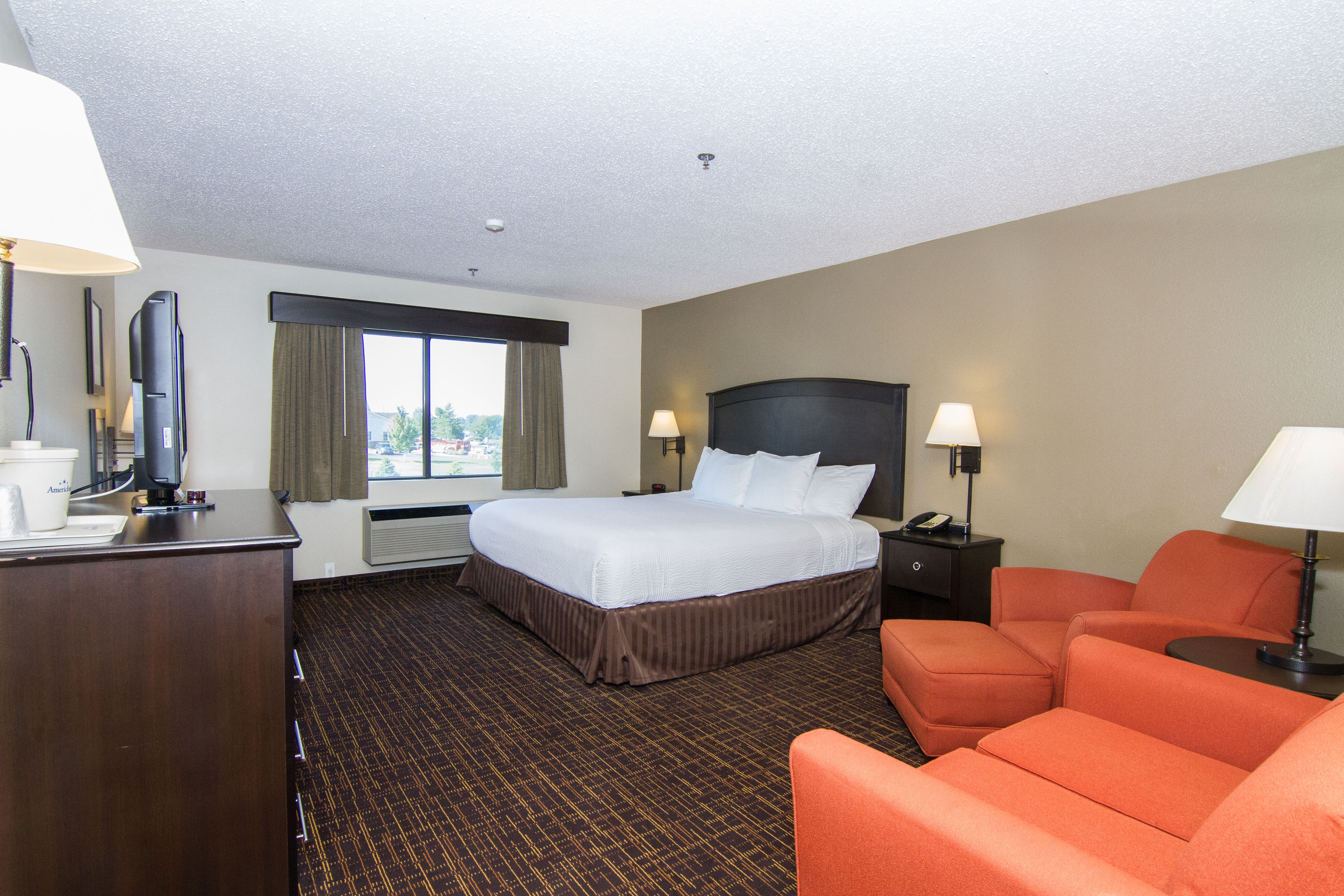 A hotel room with a large bed, two bedside tables with lamps, a desk with a computer, and a window with curtains.