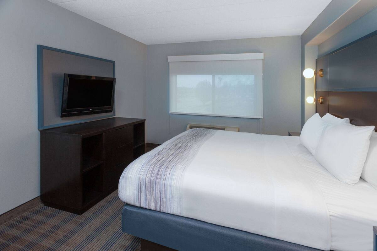 A hotel room with a large bed, a flat-screen TV mounted on the wall, a window with blinds, and a nightstand with a lamp.