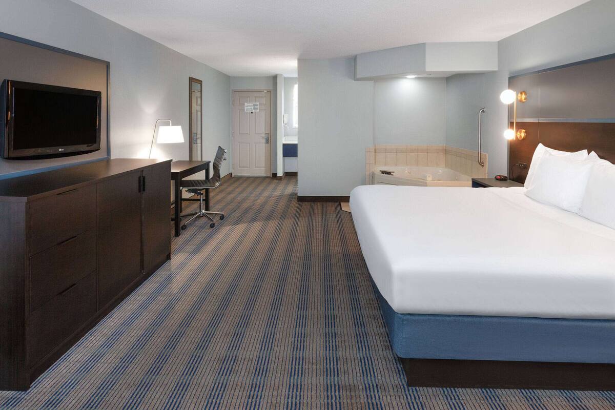 A hotel room with a large bed, a desk with a chair, a television, a bathroom with a bathtub, and a lighted wall mirror.