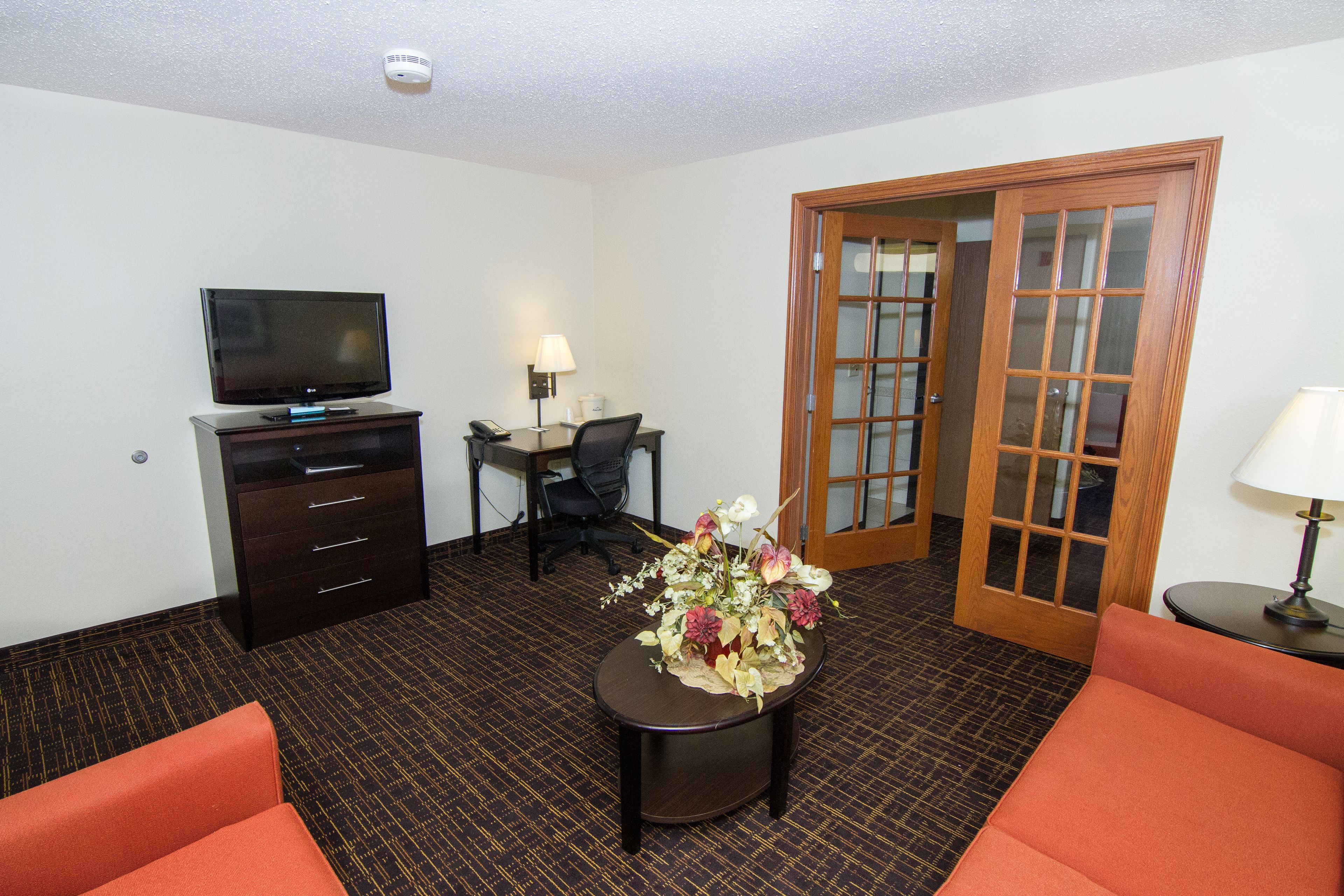 Quarto de hotel com televisão, escrivaninha com luminária, cadeira, arranjo floral e dois sofás laranja.