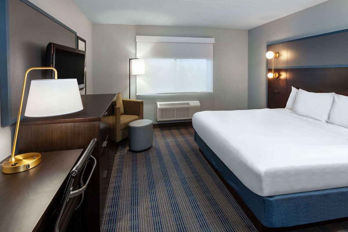 A hotel room with a bed, desk, chair, lamp, TV, and air conditioning unit.