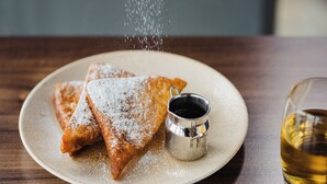 A plate with two triangular pastries dusted with powdered sugar, a small metal cup with a dark liquid, and a glass of amber liquid on a wooden table. - Carlton George Hotel (Glasgow)