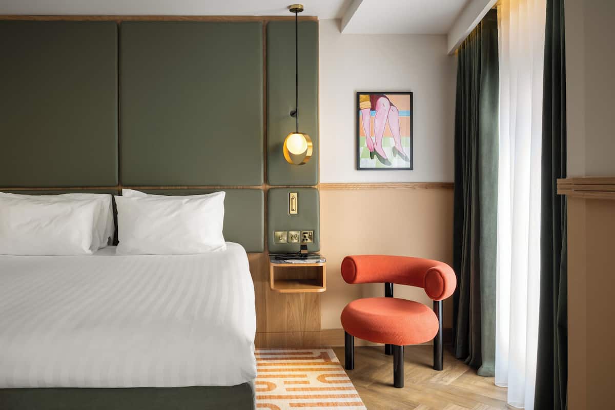 A hotel room with a large bed, a small wooden nightstand, a modern orange chair, and a painting on the wall.