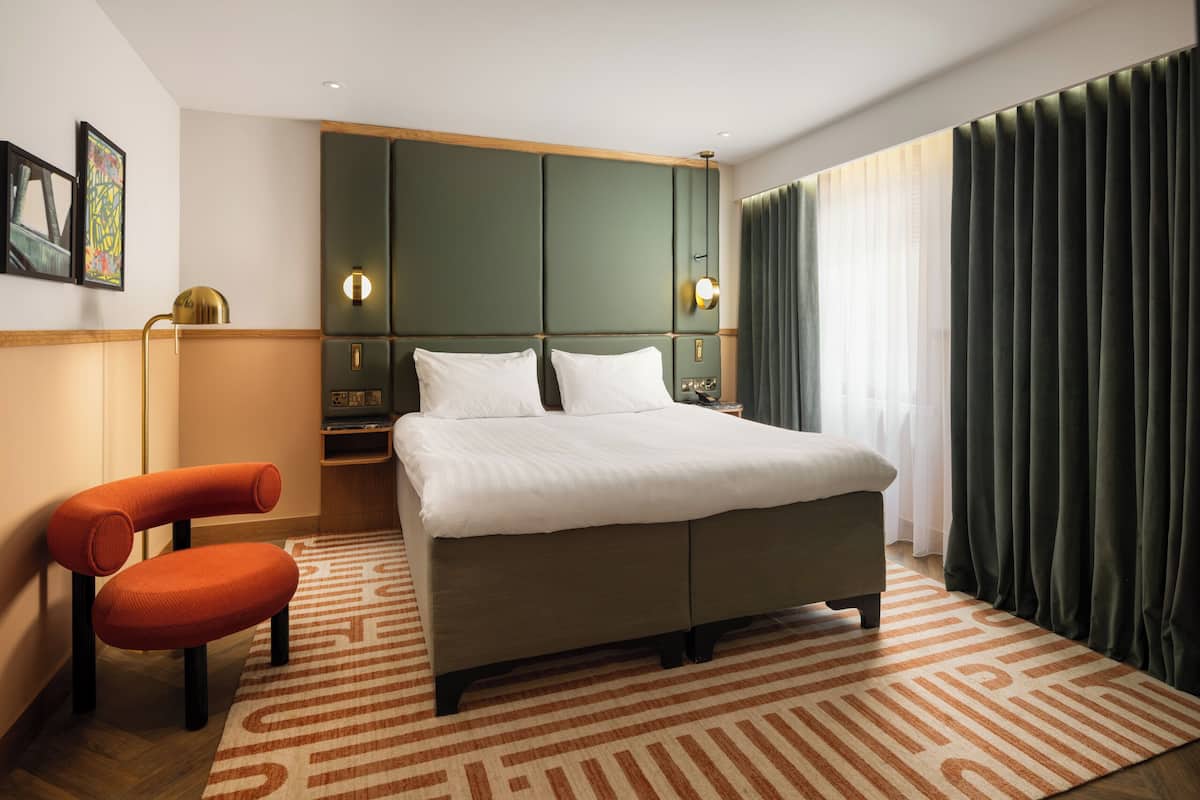A modern hotel room with a large bed, a seating area with a chair, a side table, and a wall-mounted lamp.