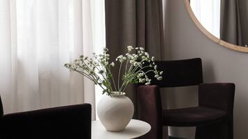 A modern room with a round mirror, two dark chairs, a small white table with a vase of flowers, and a window with sheer curtains.