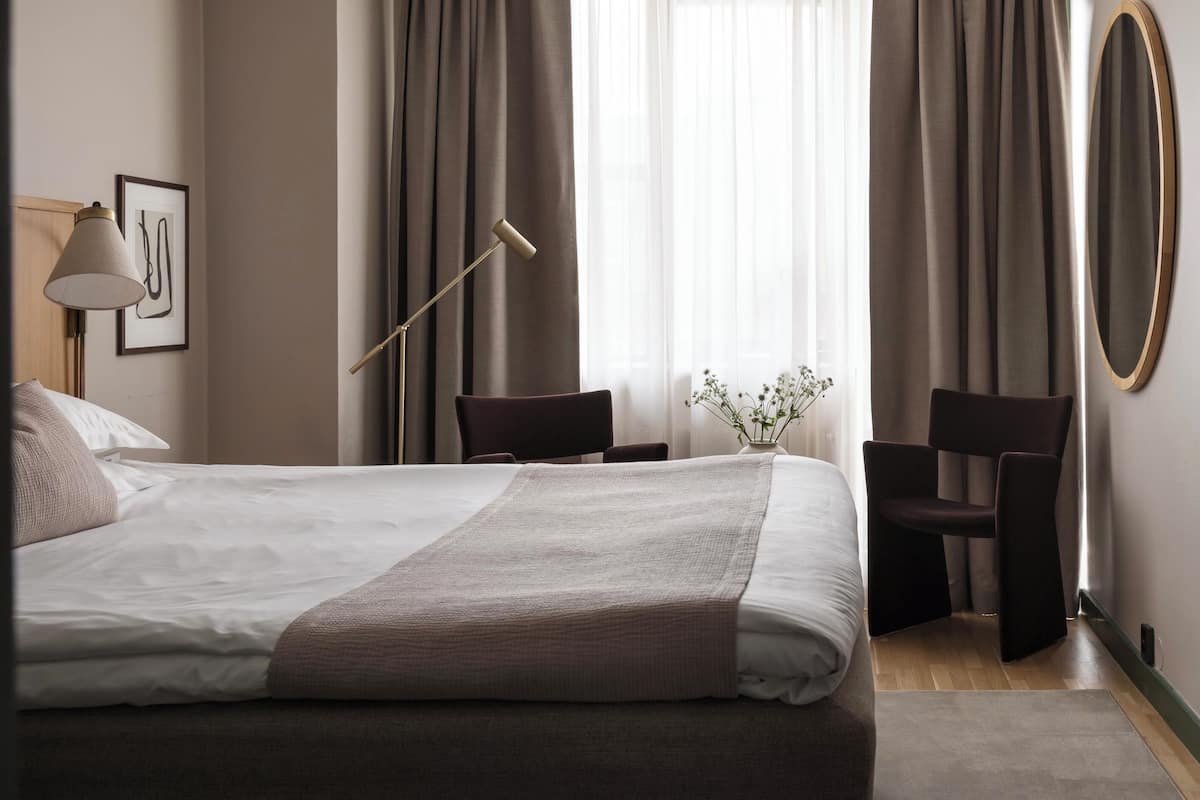 A neatly made bed with a white and brown blanket, two dark chairs, a floor lamp, and a vase with flowers on a wooden floor.