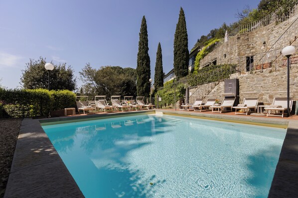 A swimming pool with lounge chairs, tall cypress trees, and stone walls.