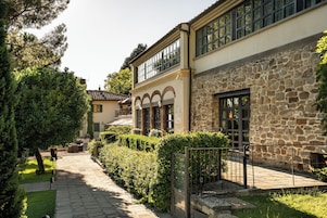 A courtyard with a stone building, a paved walkway, and a hedge.