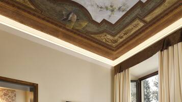 A room with a high ceiling featuring a detailed fresco, a large window with curtains, and a framed mirror on the wall.