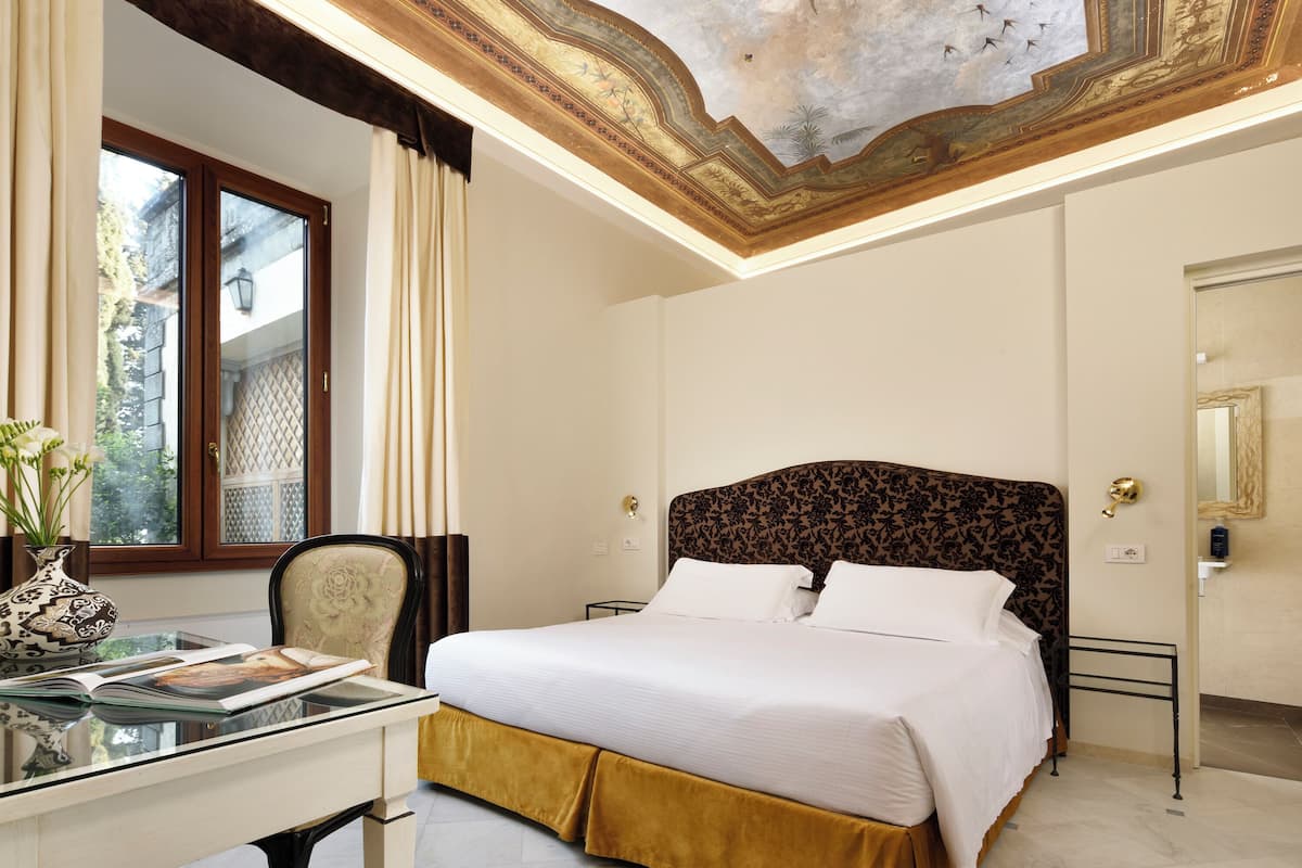Habitación de hotel con una cama grande, un escritorio con un florero y un techo con detalles artísticos.