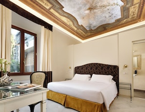 A hotel room with a large bed, a desk with a vase, and a ceiling with detailed artwork.