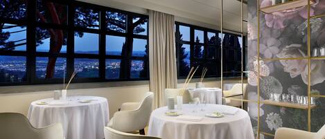 A dining area with round tables, white tablecloths, and chairs, featuring a large window with a city view at dusk.