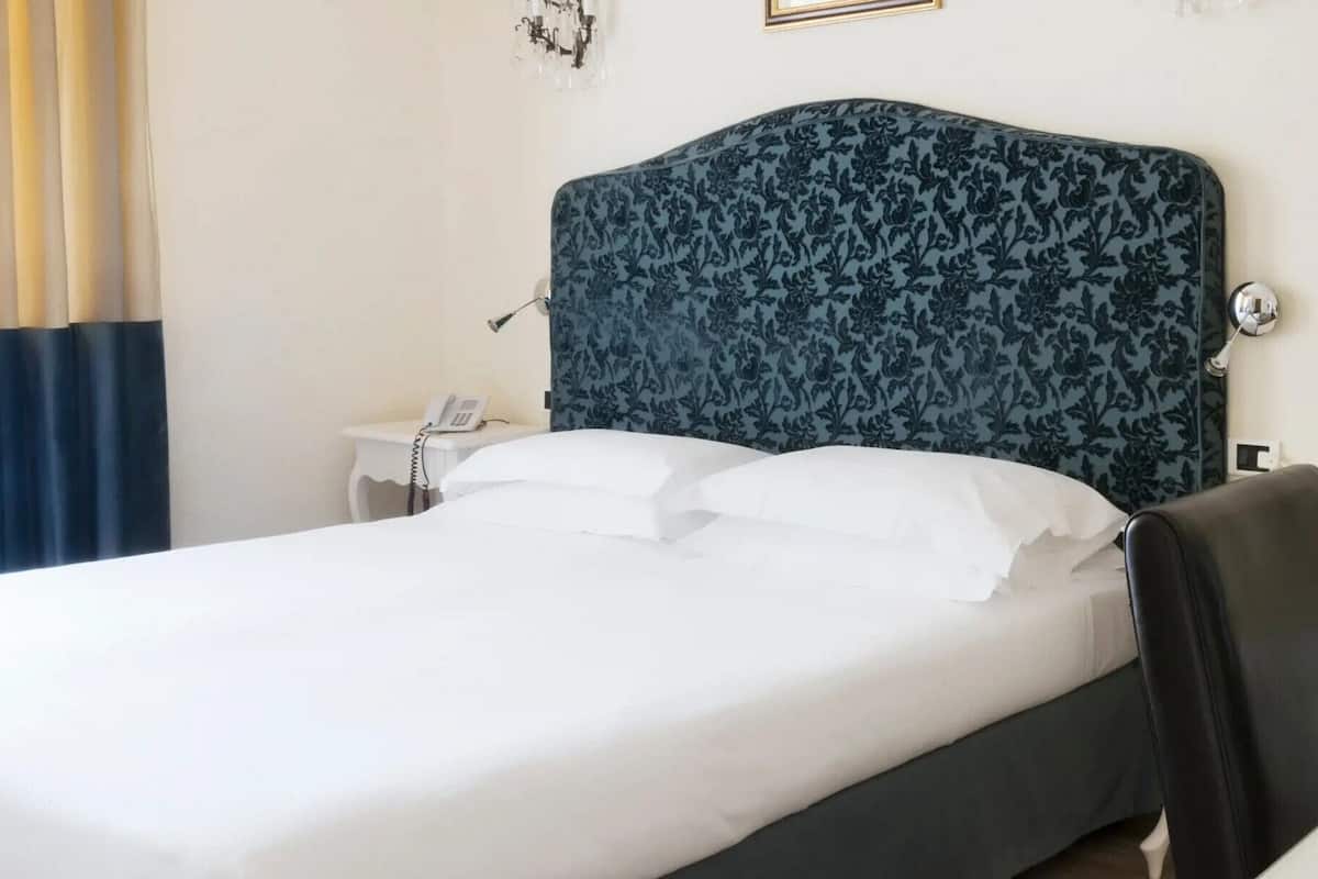Una cama bien hecha con sábanas blancas, un cabecero azul oscuro con motivos florales, un teléfono en la mesita de noche y una pequeña mesa con una lámpara.
