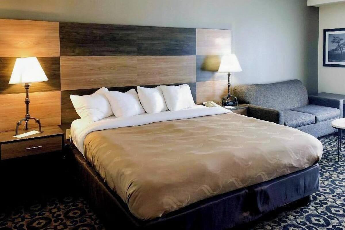 A hotel room with a large bed, two bedside lamps, a grey sofa, and a small round table.