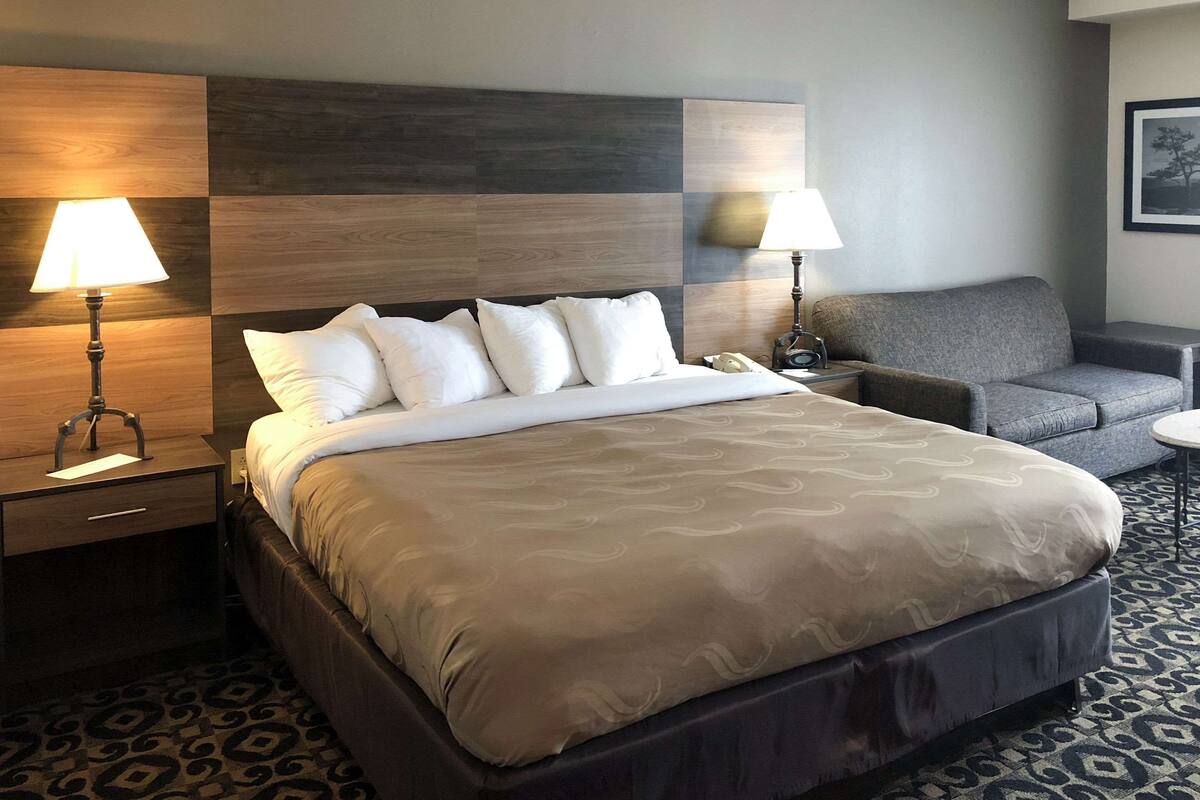 A hotel room with a large bed, two bedside lamps, a gray sofa, and a patterned carpet.