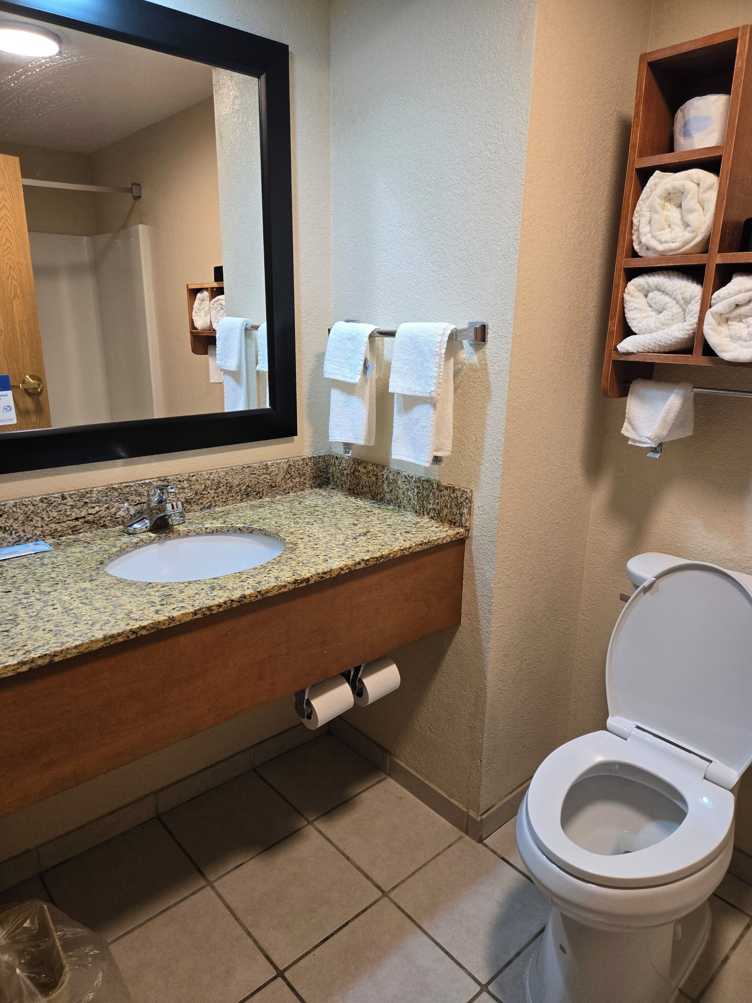 A bathroom with a granite countertop sink, a mirror, a toilet, and a towel rack with folded towels.