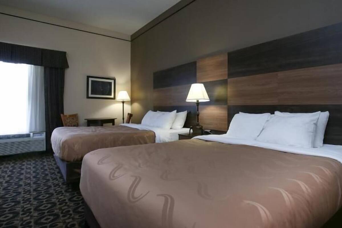 A hotel room with two beds, a desk, a chair, a lamp, and a framed picture on the wall.