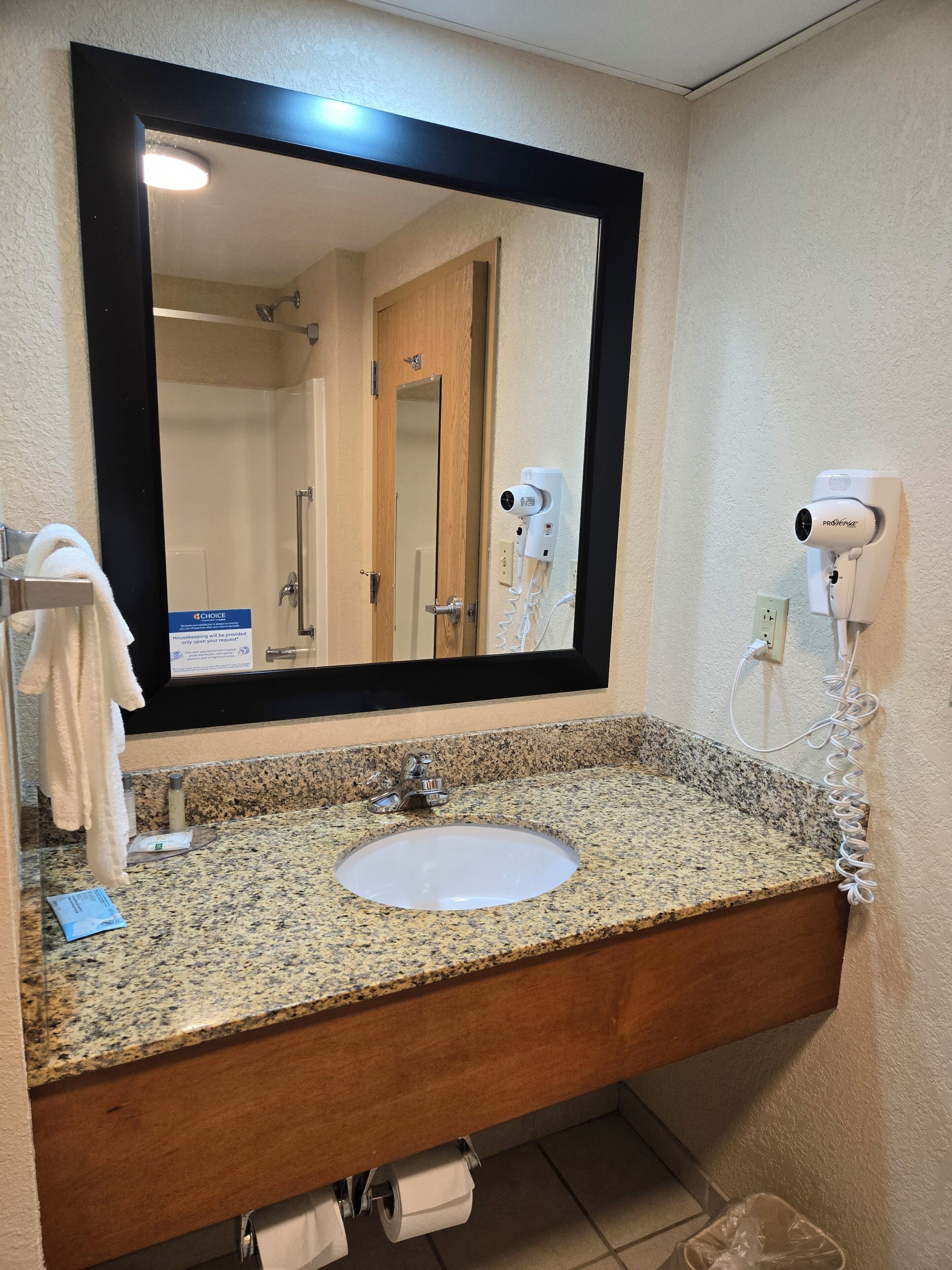 A bathroom with a granite countertop sink, a mirror with a black frame, a towel rack, a hairdryer, and a sign on the wall.