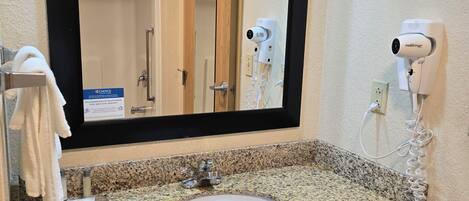 A bathroom with a granite countertop sink, a mirror with a black frame, a towel rack, a hairdryer, and a sign on the wall.