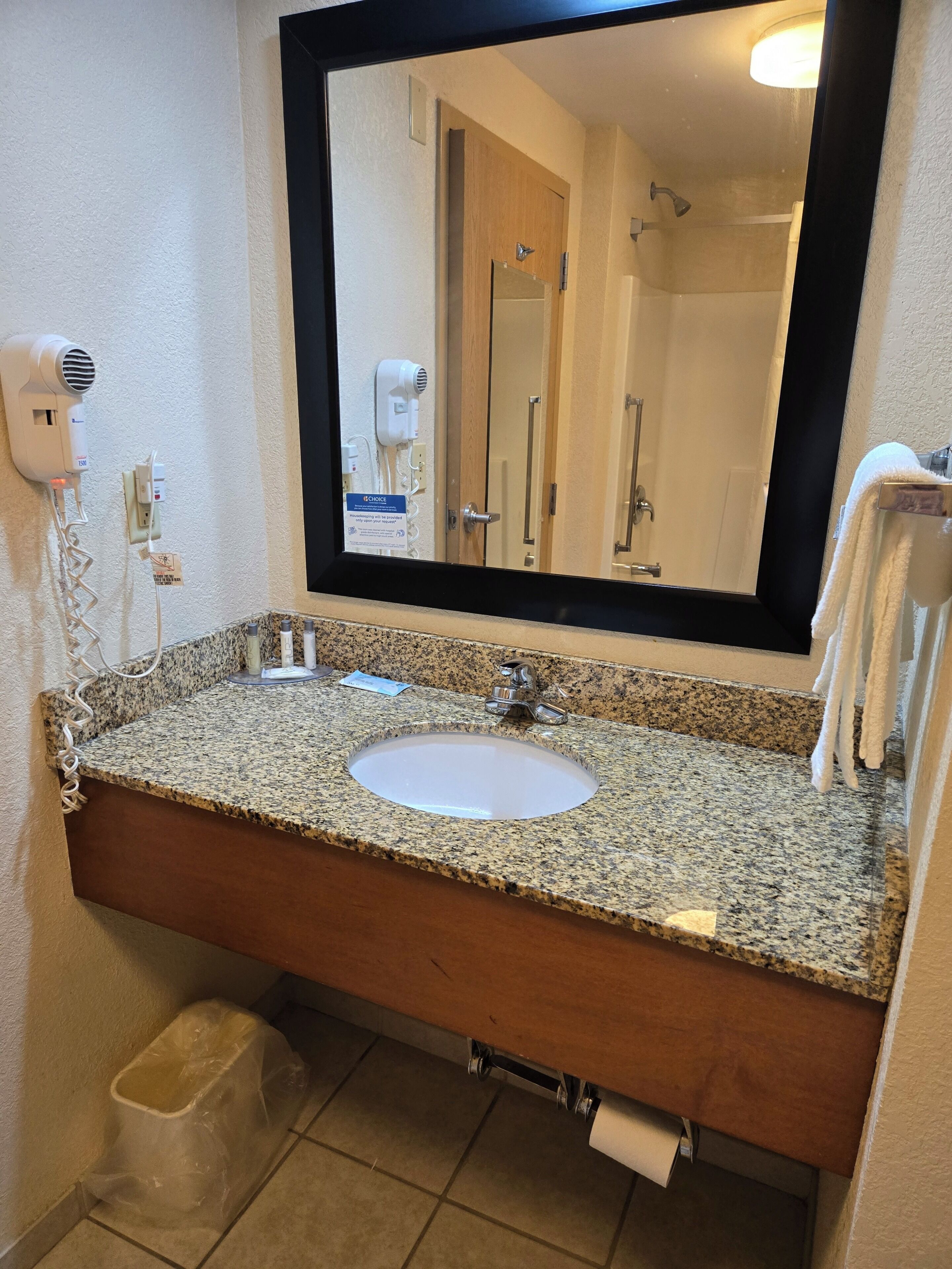 Un baño con lavamanos de encimera de granito, un espejo, toallero, secador de pelo y un cubo de basura.