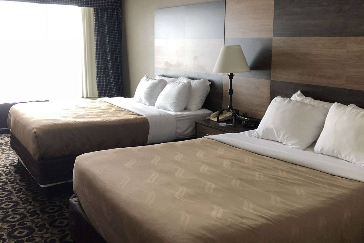A hotel room with two beds, a nightstand with a lamp, and a large window with curtains.