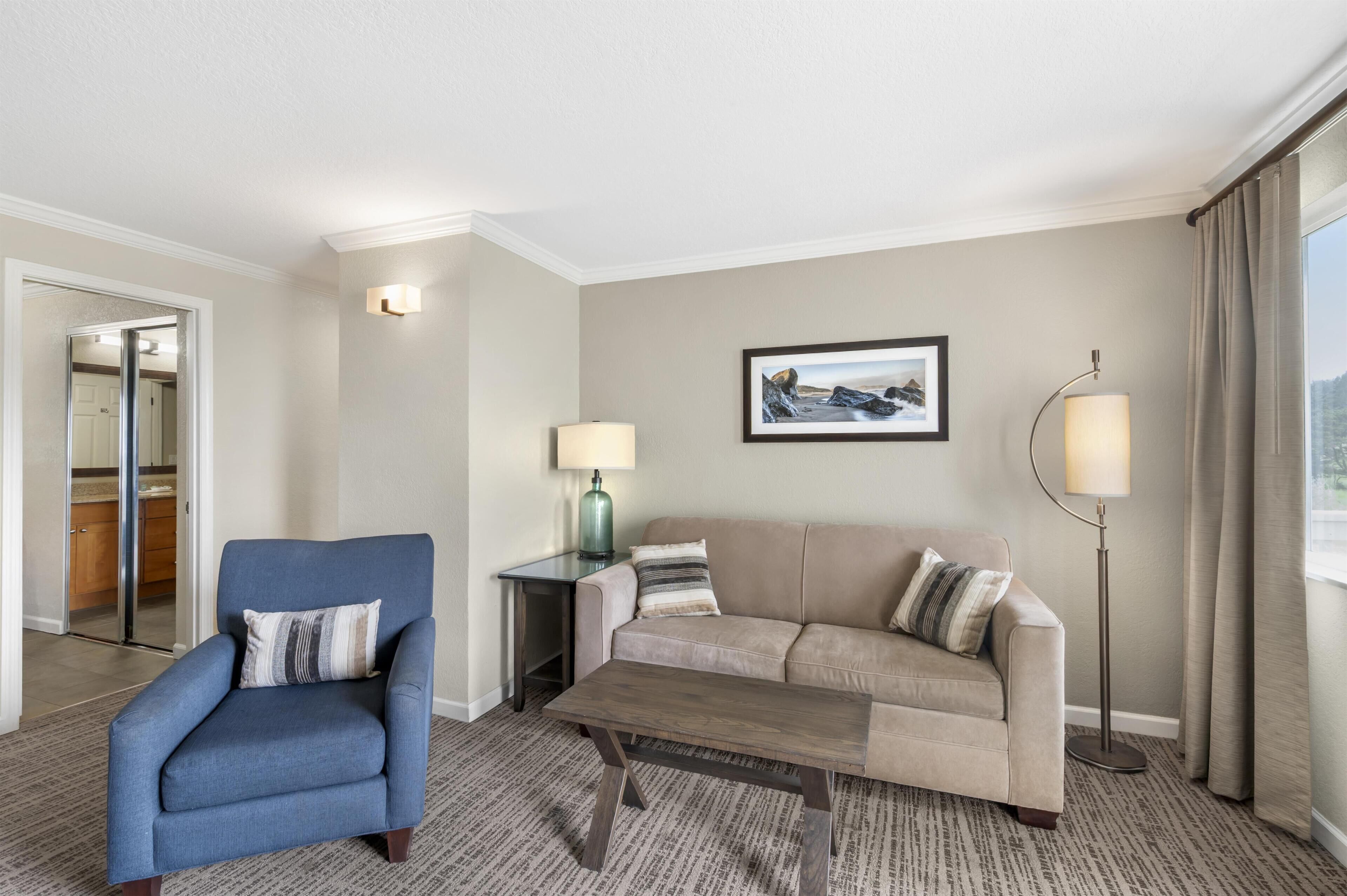 A hotel room with a blue armchair, a beige sofa, a coffee table, a side table with a lamp, a framed picture on the wall, and a view of the outdoors through a window.