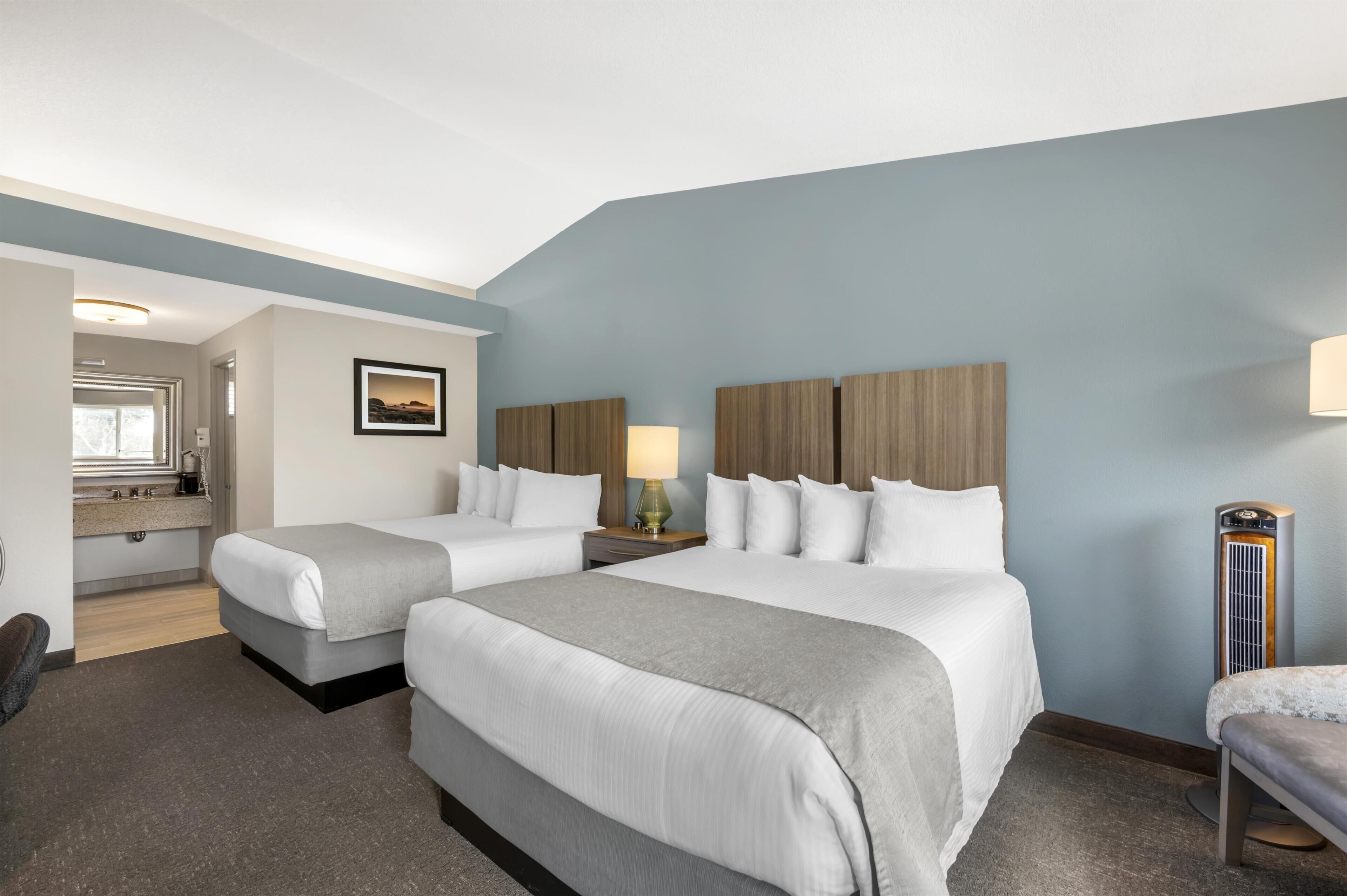 A hotel room with two beds, a bedside table with a lamp, a large mirror, and a view of the hallway.