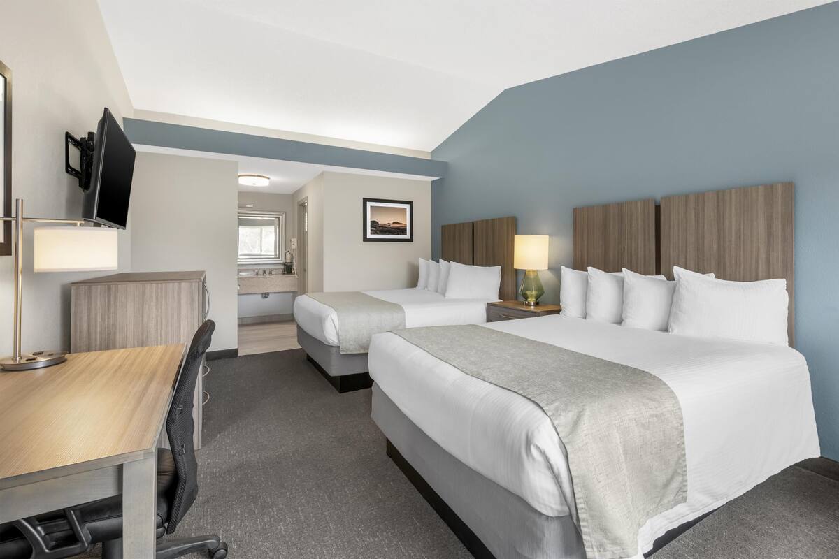 Habitación de hotel con dos camas, un escritorio, una silla, un televisor y un baño visible a través de una puerta abierta.