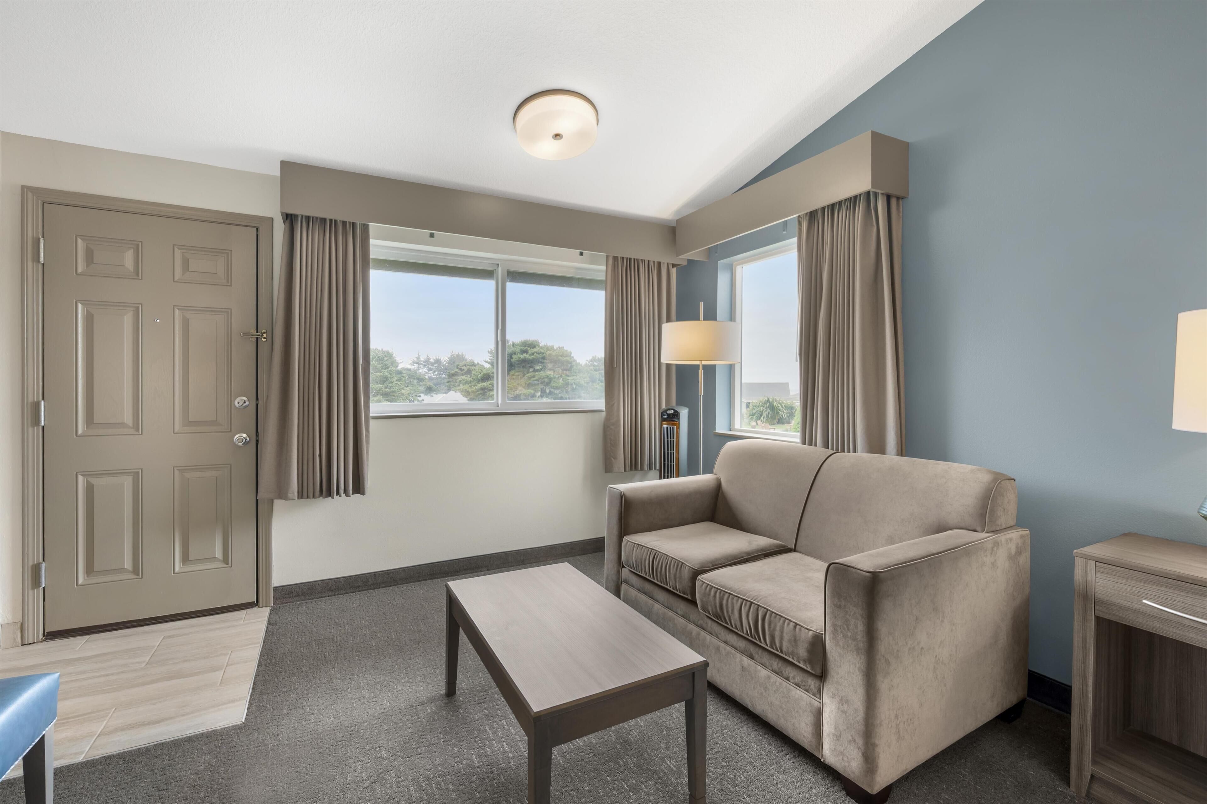Ein Hotelzimmer mit einer Couch, einem kleinen Tisch, einer Lampe, einer Tür und einem Fenster mit Vorhängen.