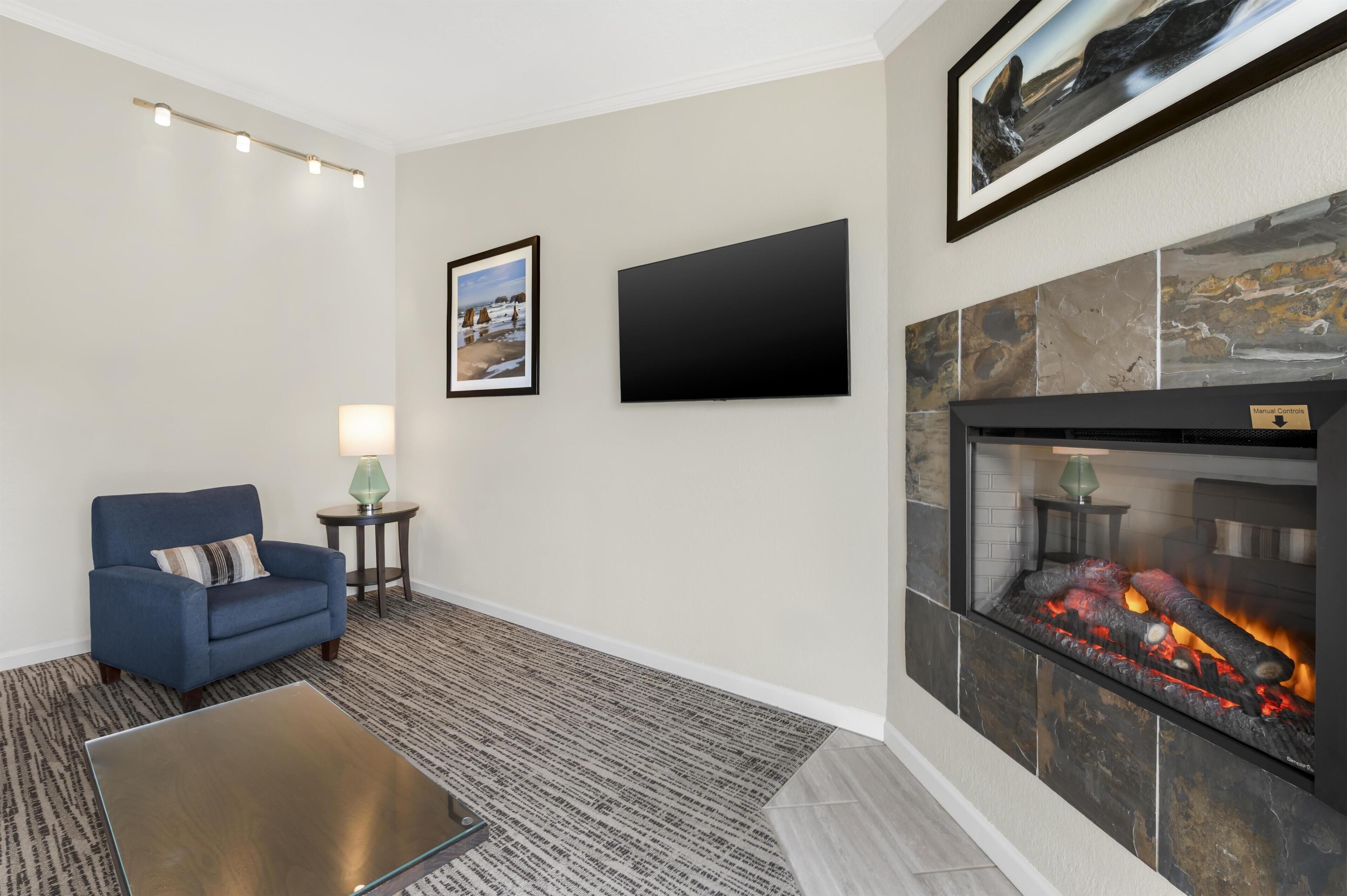 A living room with a blue armchair, a glass coffee table, a fireplace with a fire, a flat-screen TV, and a framed picture on the wall.