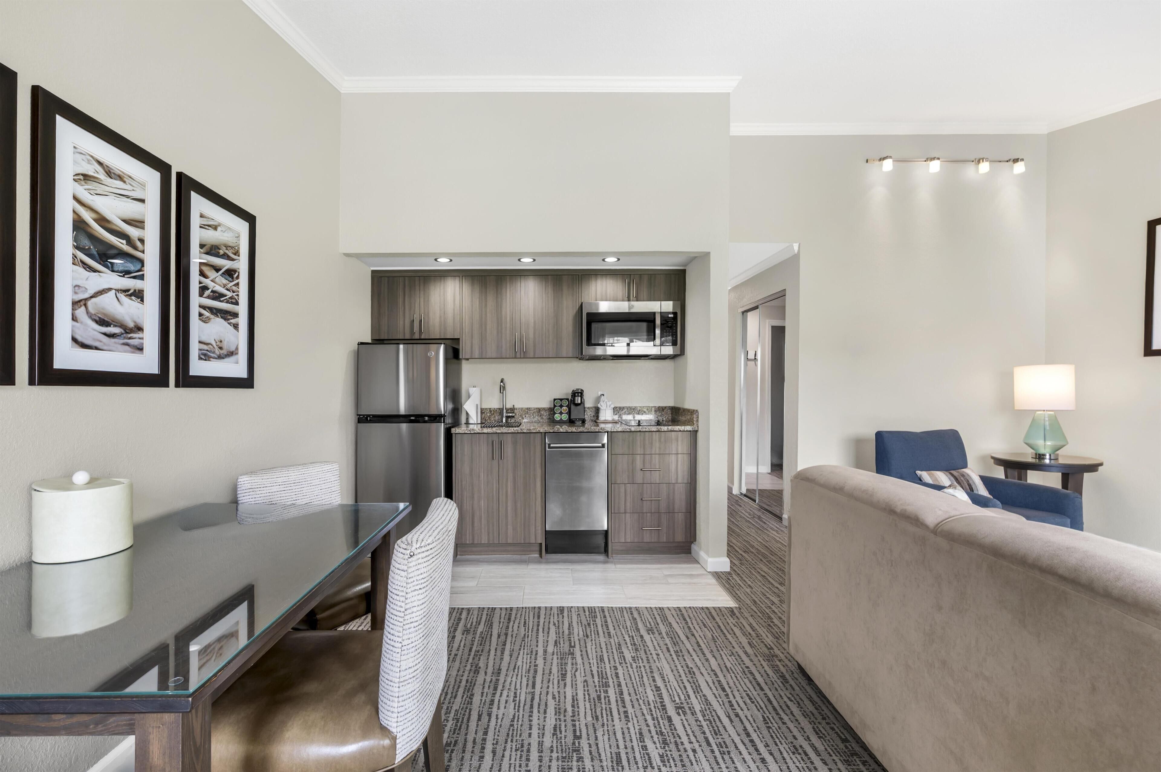 A modern hotel room with a kitchenette, a dining area, a sofa, and a small table with a lamp.